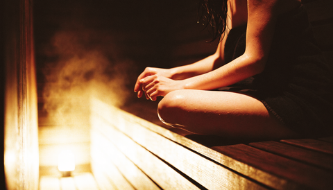 Sauna și longevitatea: o femeie stă pe banca de lemn a unei saune tradiționale, în lumină caldă și aburi.