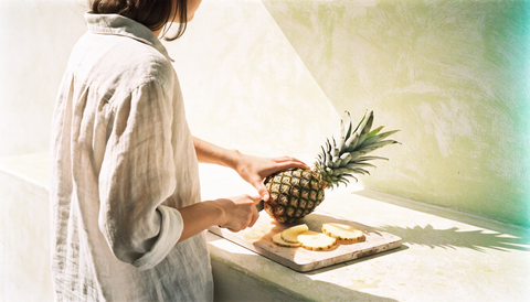Bromelaina și papaina: o femeie taie un ananas pe o lespede de piatră palidă, cu fața întoarsă, în lumina care aruncă o umbră lungă pe peretele de tencuială.